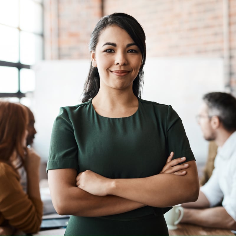 A woman with dark hair wearing a dark green shirt smiling with her arms crossed, standing in front of a group of people at a conference room table
